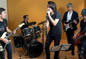 Una banda de 5 jóvenes tocando varios instrumentos: guitarra, bajo, batería. En el centro uno de ellos canta con un micrófono en el Estudio Samela de Educación Musical (ESSEM)